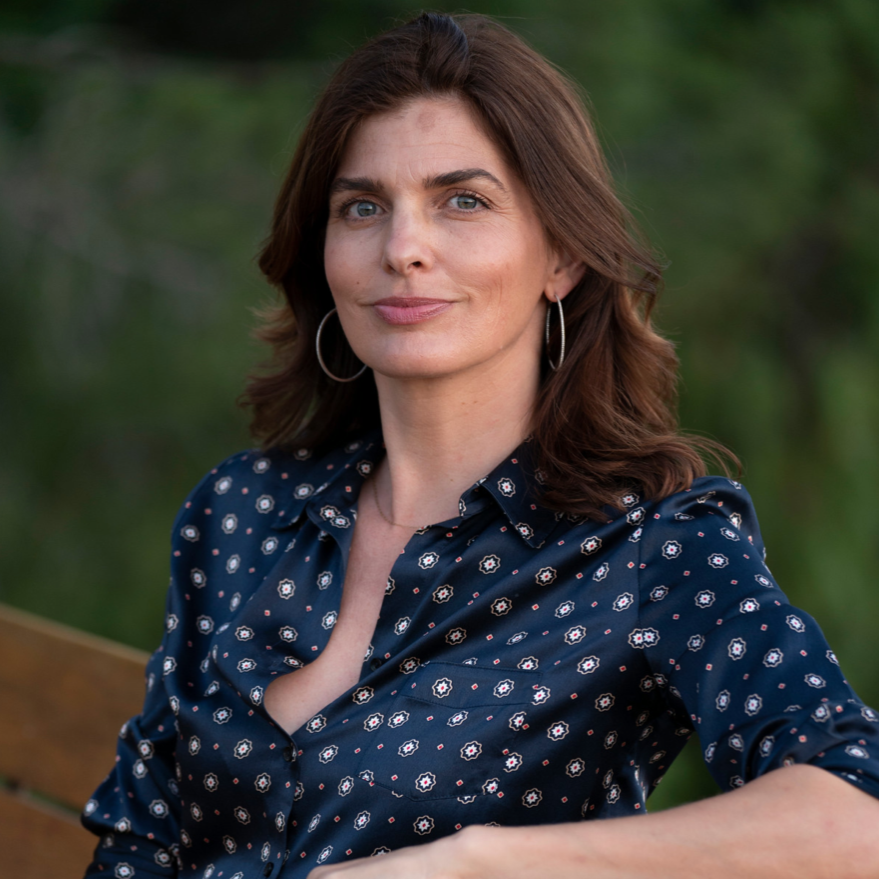 Sitting on a wooden bench wearing a polka dot shirt and jeans, with a blurred background.